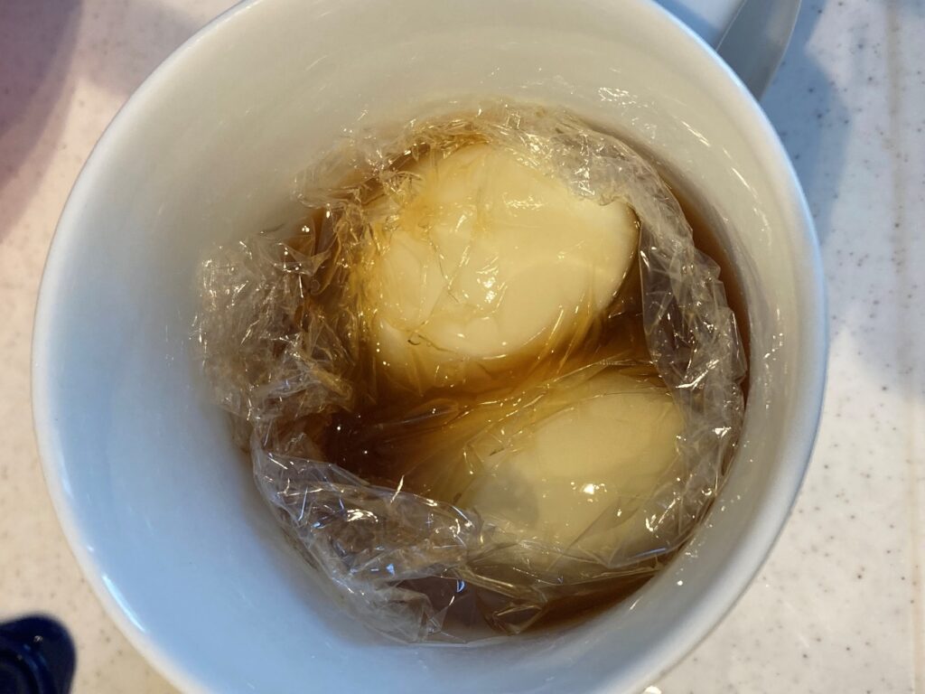 マグカップに業務スーパーの鶏照り焼きの素と水を入れ向いたゆで卵を入れラップで落とし蓋